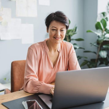 Middle aged woman smiles and uses laptop in home office at desk